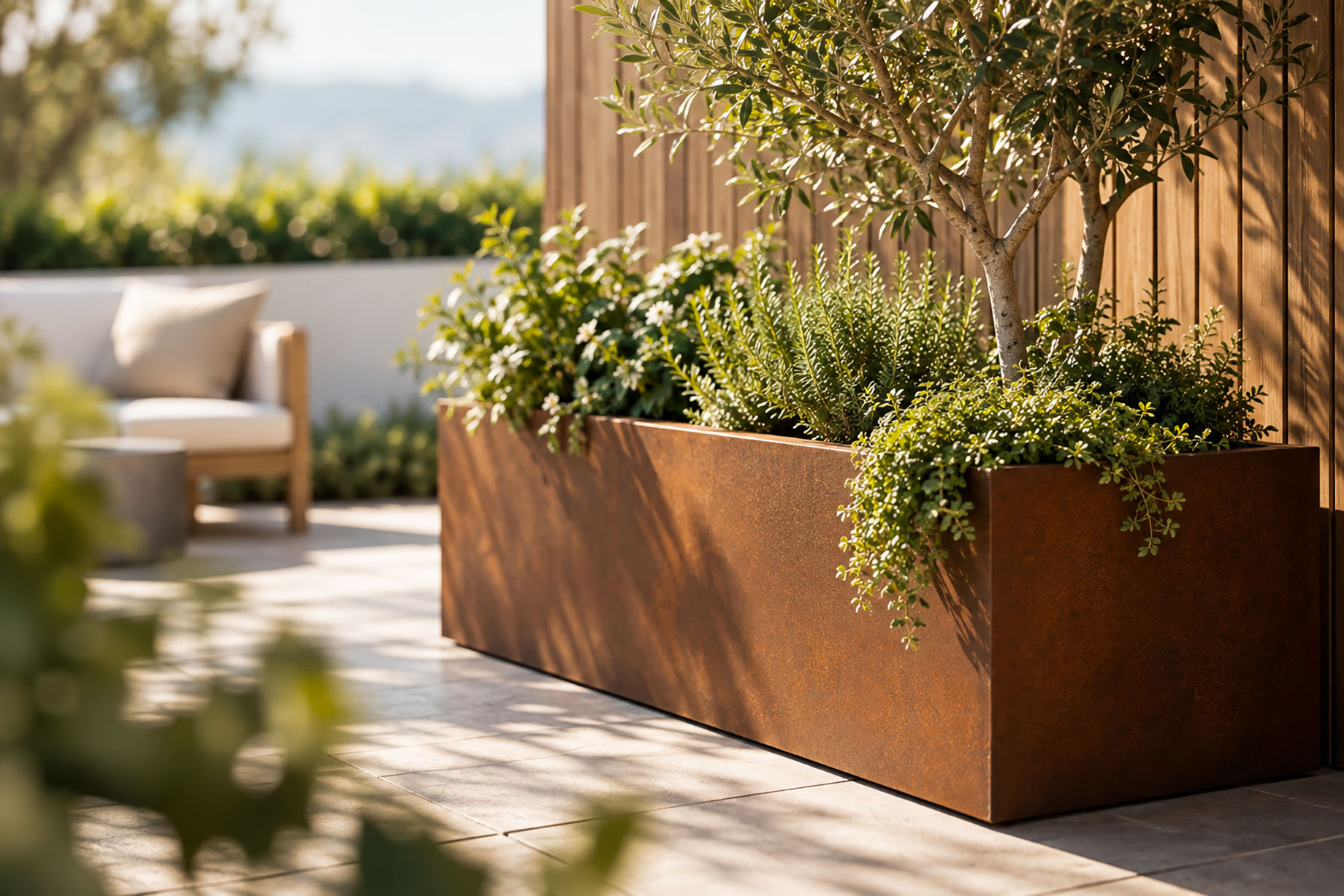Large corten steel planter box on a sunlit patio with a small tree, rosemary, trailing greenery, and flowers against a timber wall.