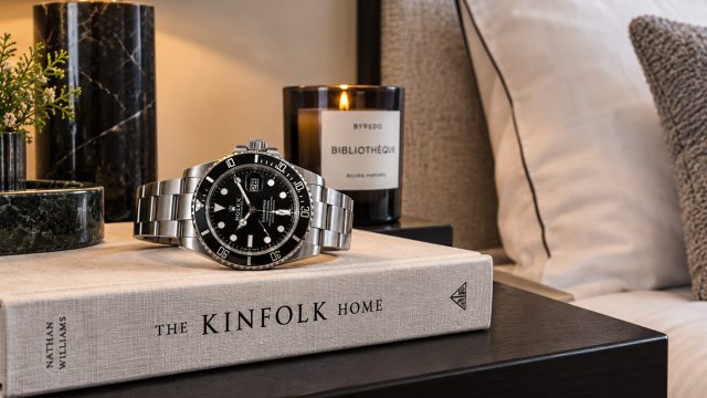 Steel luxury sports watch resting on a neutral hardback beside a marble lamp and candle in an elegantly staged bedroom.