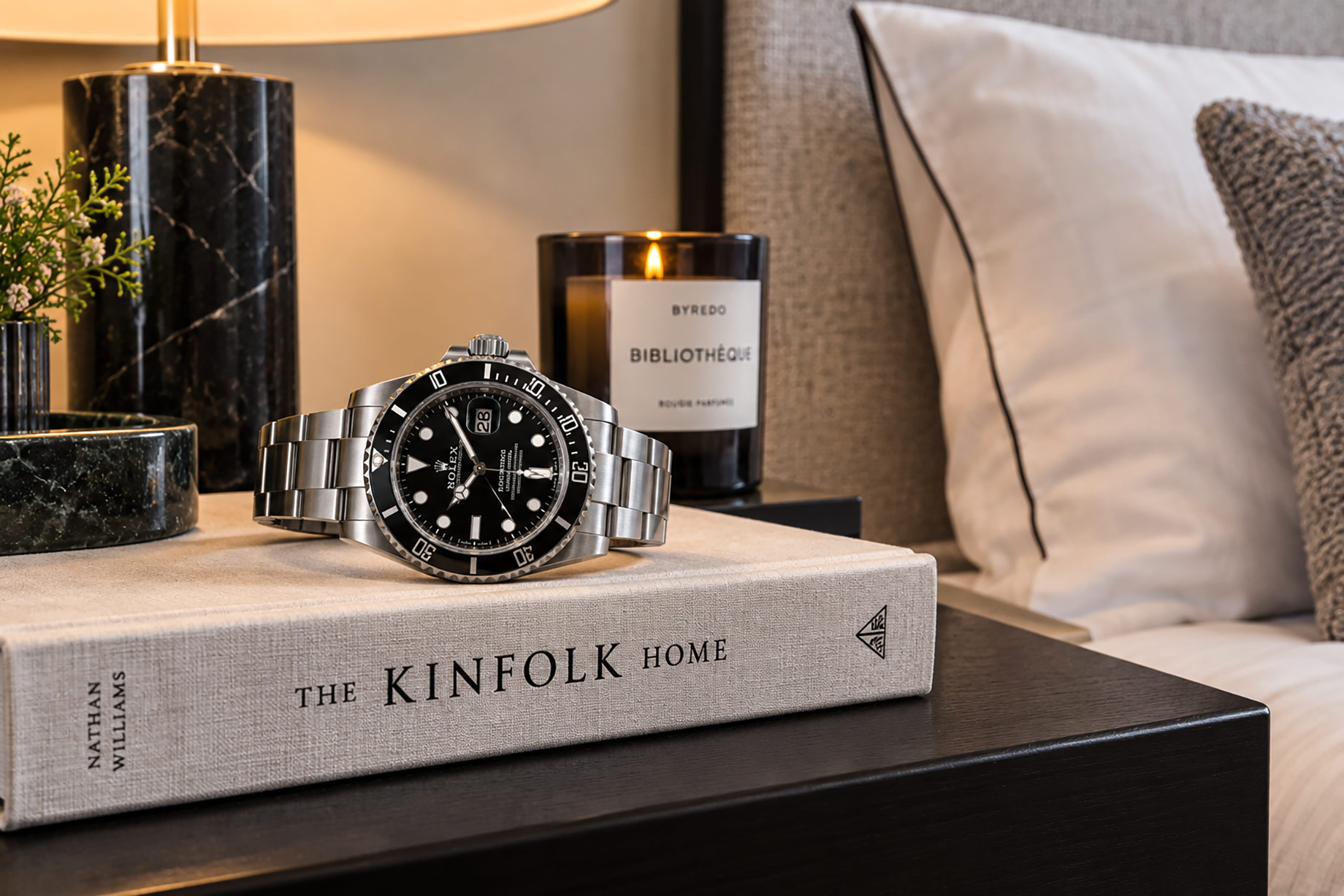 Steel luxury sports watch resting on a neutral hardback beside a marble lamp and candle in an elegantly staged bedroom.
