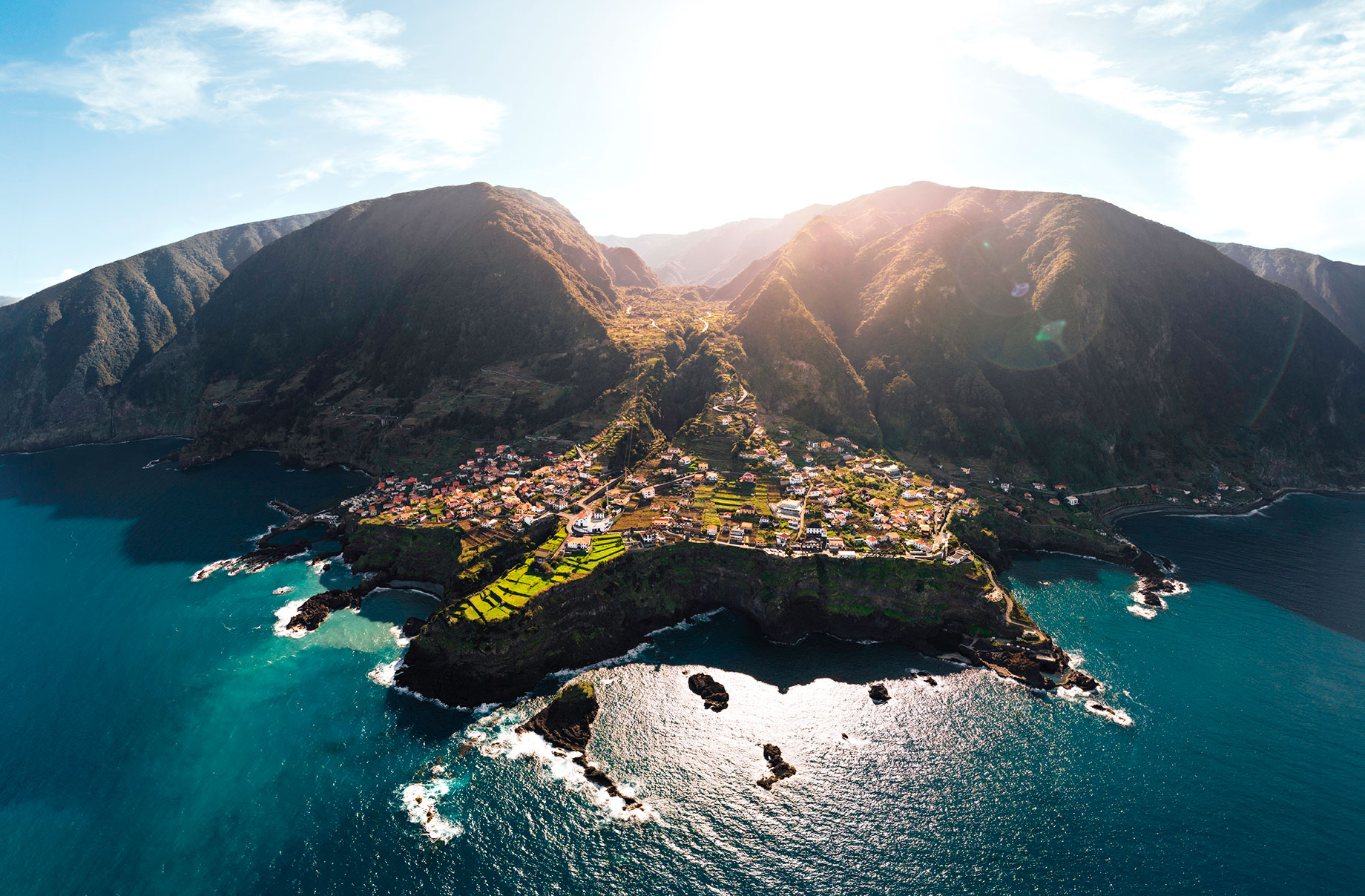 Coastal village on Madeira with red-roofed homes, green terraces, rocky cliffs, and steep mountains above the Atlantic.