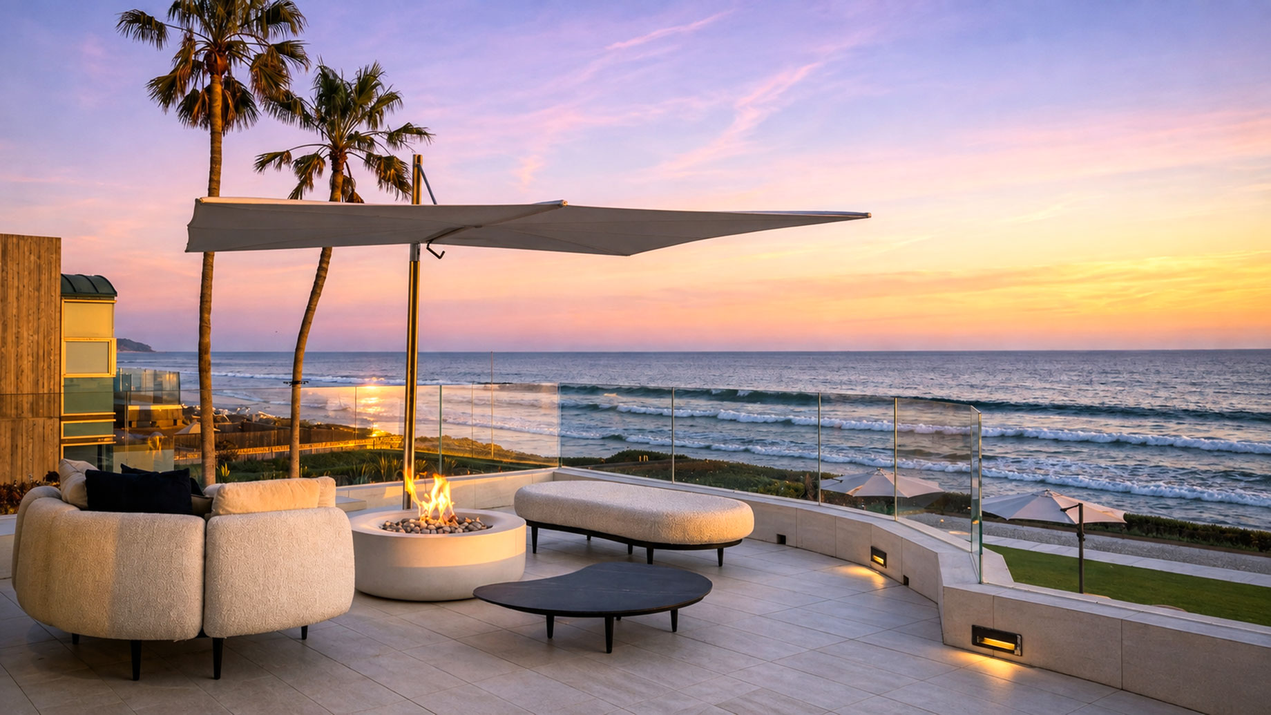 Cream seating and a fire pit beneath a cantilever umbrella on a Malibu terrace with glass railings and ocean views at sunset.
