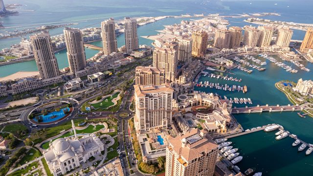 Aerial view of The Pearl Island in Doha, showing Porto Arabia’s waterfront towers, marina, yachts, landscaped roads, and turquoise Gulf waters.
