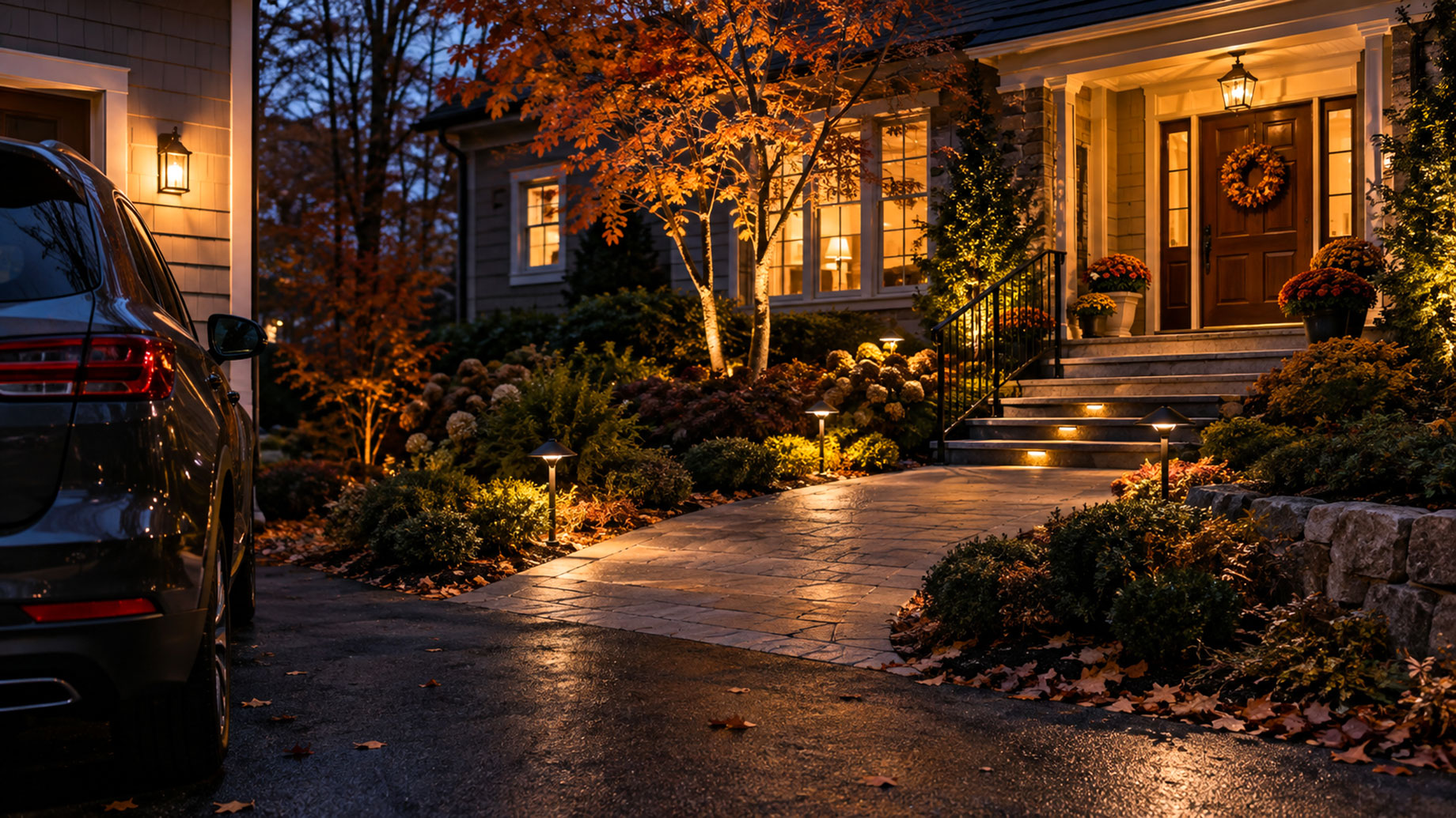 Illuminated walkway leading from a parked car to a front entrance with visible steps, handrails, path lights, and autumn landscaping.