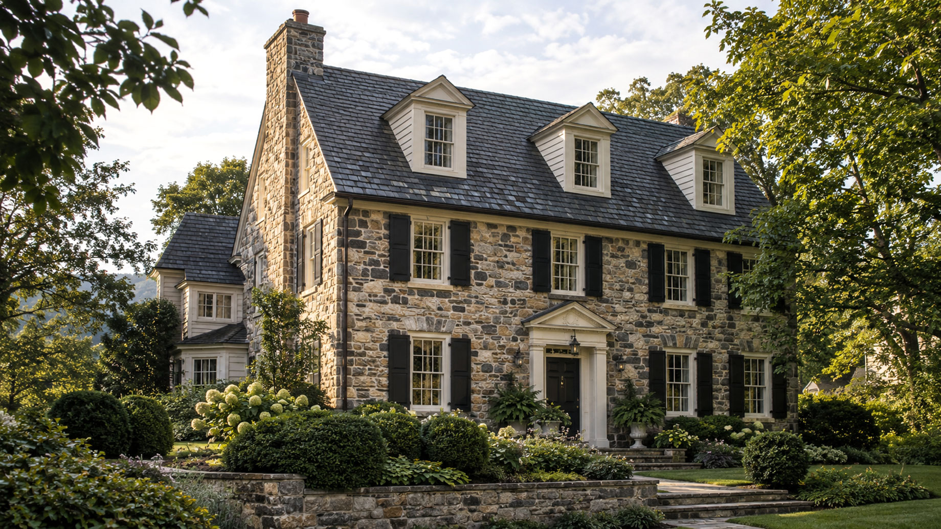 Stone house with a steep slate-style roof, three white dormer windows, dark shutters, and a tall chimney, framed by mature trees.
