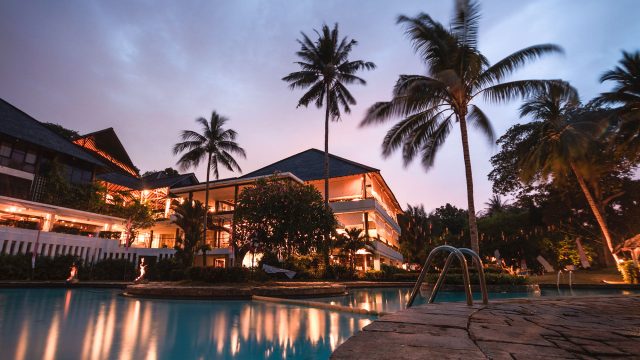 Palm trees above a tropical resort and swimming pool at dusk, with warm building lights reflected in the water beside a stone deck.