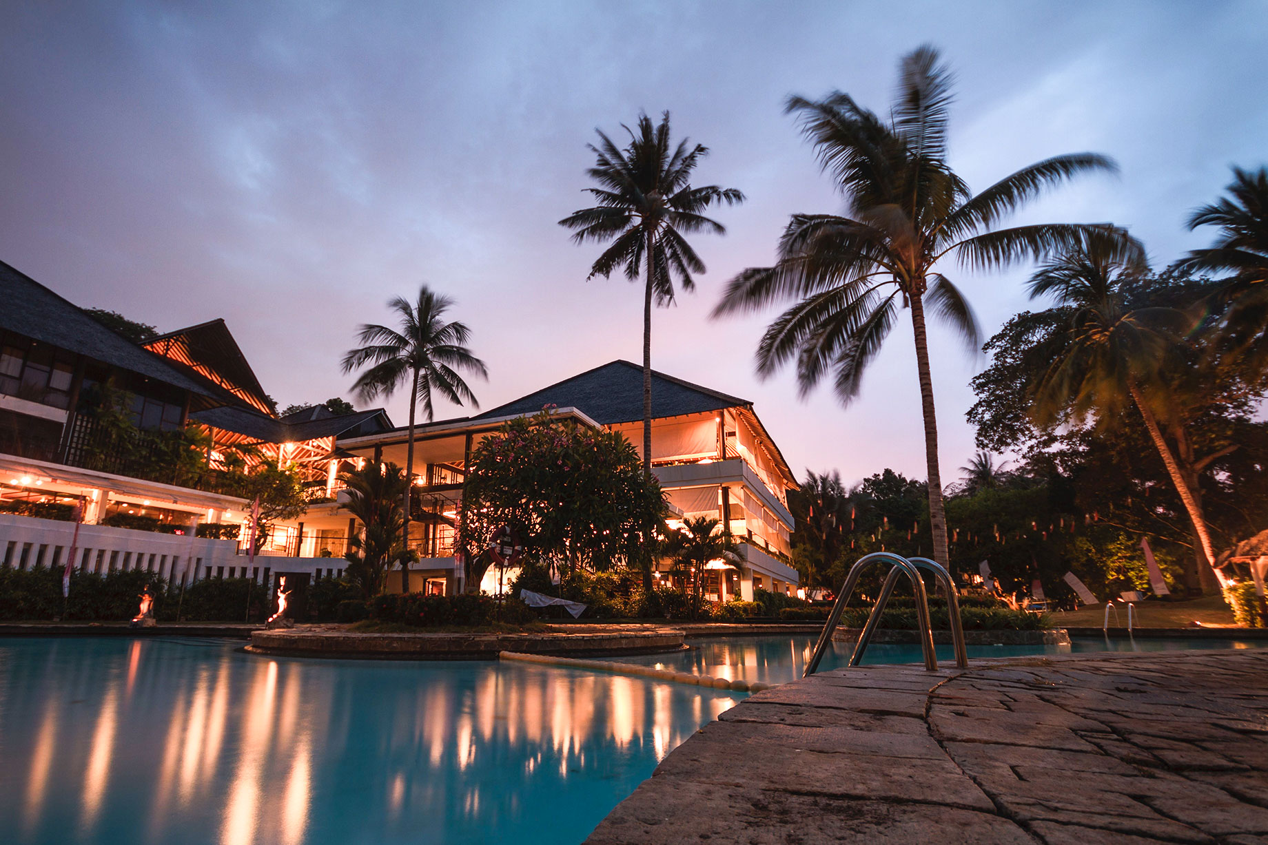 Palm trees above a tropical resort and swimming pool at dusk, with warm building lights reflected in the water beside a stone deck.
