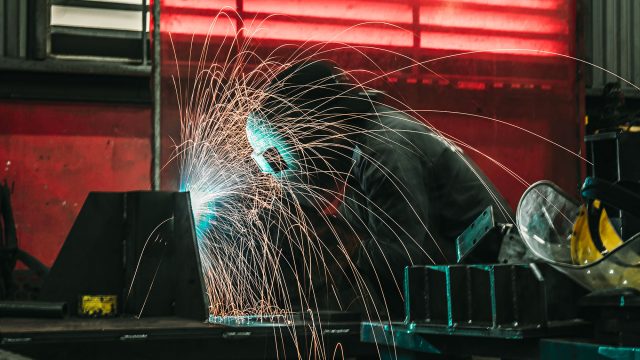 Metalworker welding a steel component in an industrial fabrication shop as bright sparks scatter across the work area.