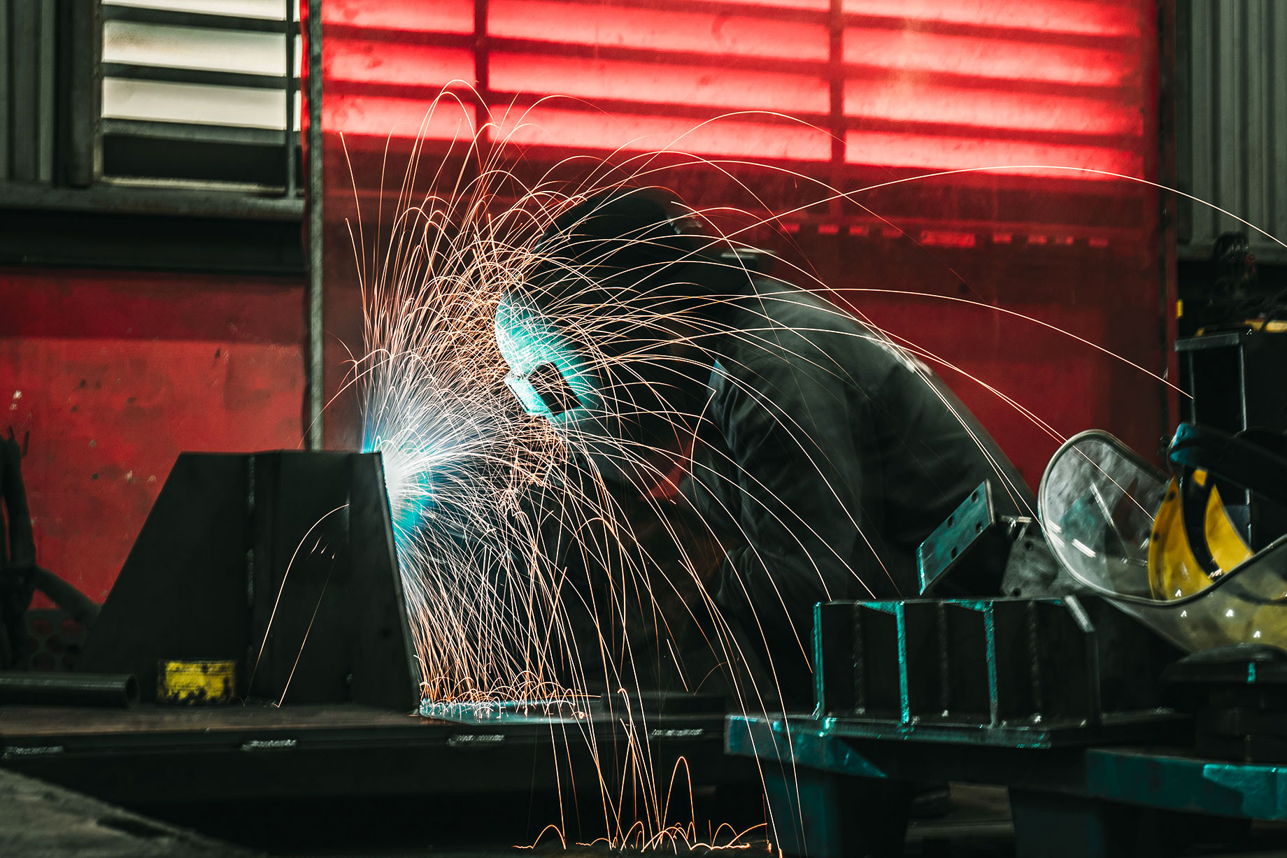 Metalworker welding a steel component in an industrial fabrication shop as bright sparks scatter across the work area.