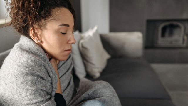 Woman sitting with her eyes closed, wrapped in a gray blanket on a sofa, with pale cushions and an unlit fireplace behind her.