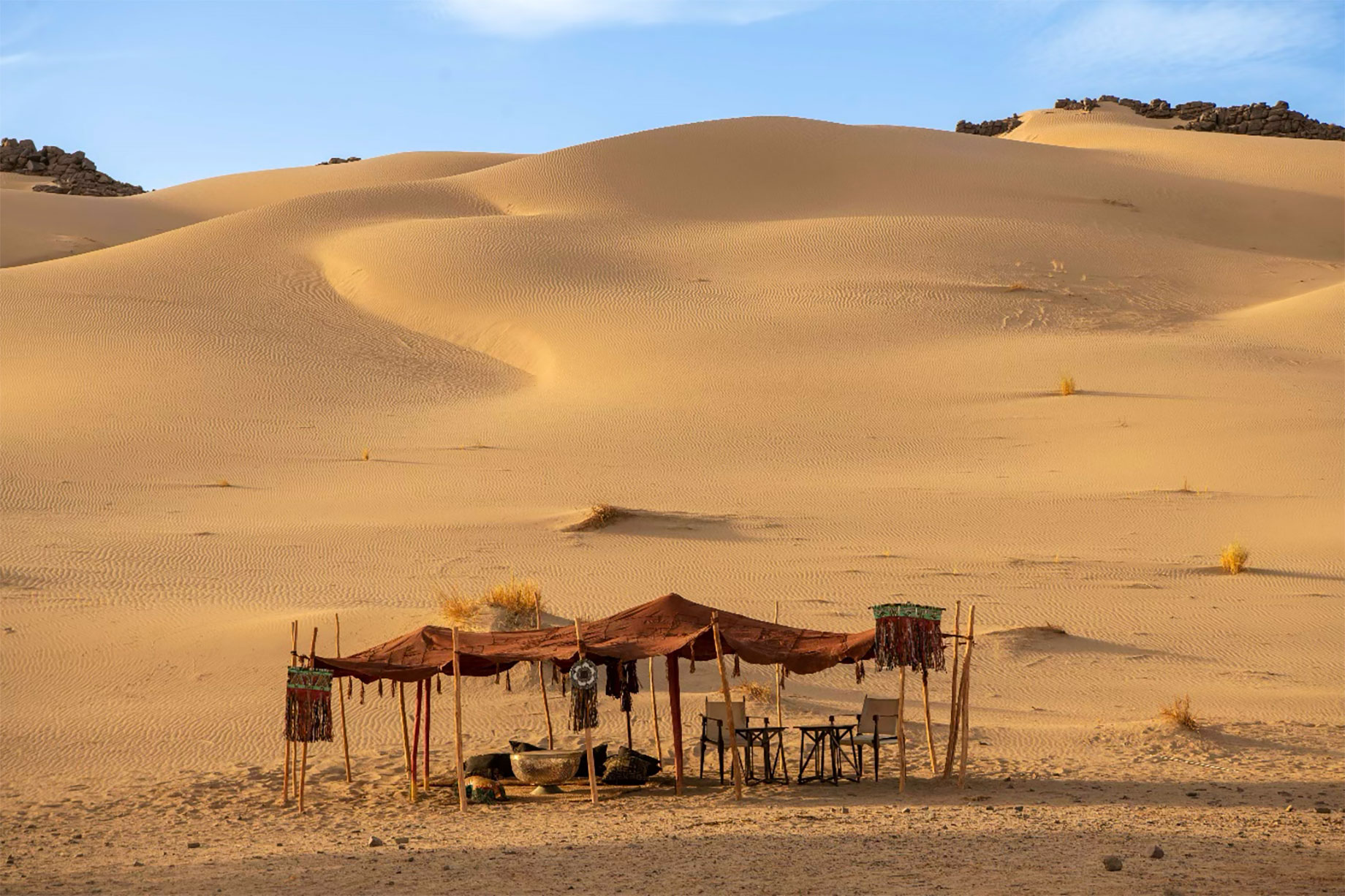 Rust-coloured canopy sheltering chairs, small tables and cushions beneath golden sand dunes and a pale blue sky.