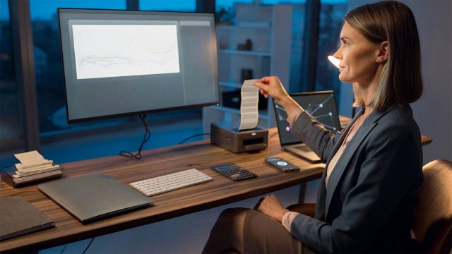 Finance professional reviewing a printed receipt beside digital financial charts, a laptop, calculator, and other accounting tools.