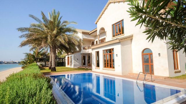 Palm Jumeirah villa with a blue swimming pool, cream facade, palm trees and landscaped gardens beside a sandy beach.
