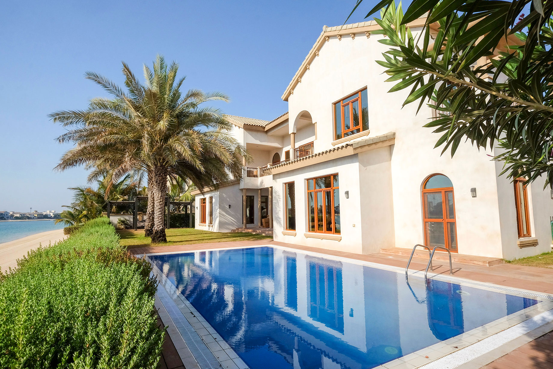 Palm Jumeirah villa with a blue swimming pool, cream facade, palm trees and landscaped gardens beside a sandy beach.