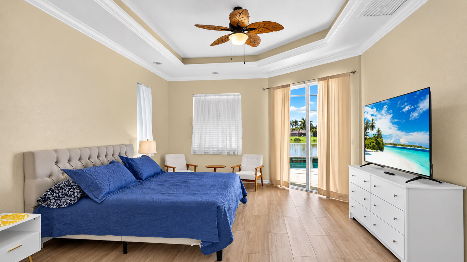 Bedroom with blue bedding, a tufted headboard, white dresser and sliding glass doors revealing pool and waterfront views.
