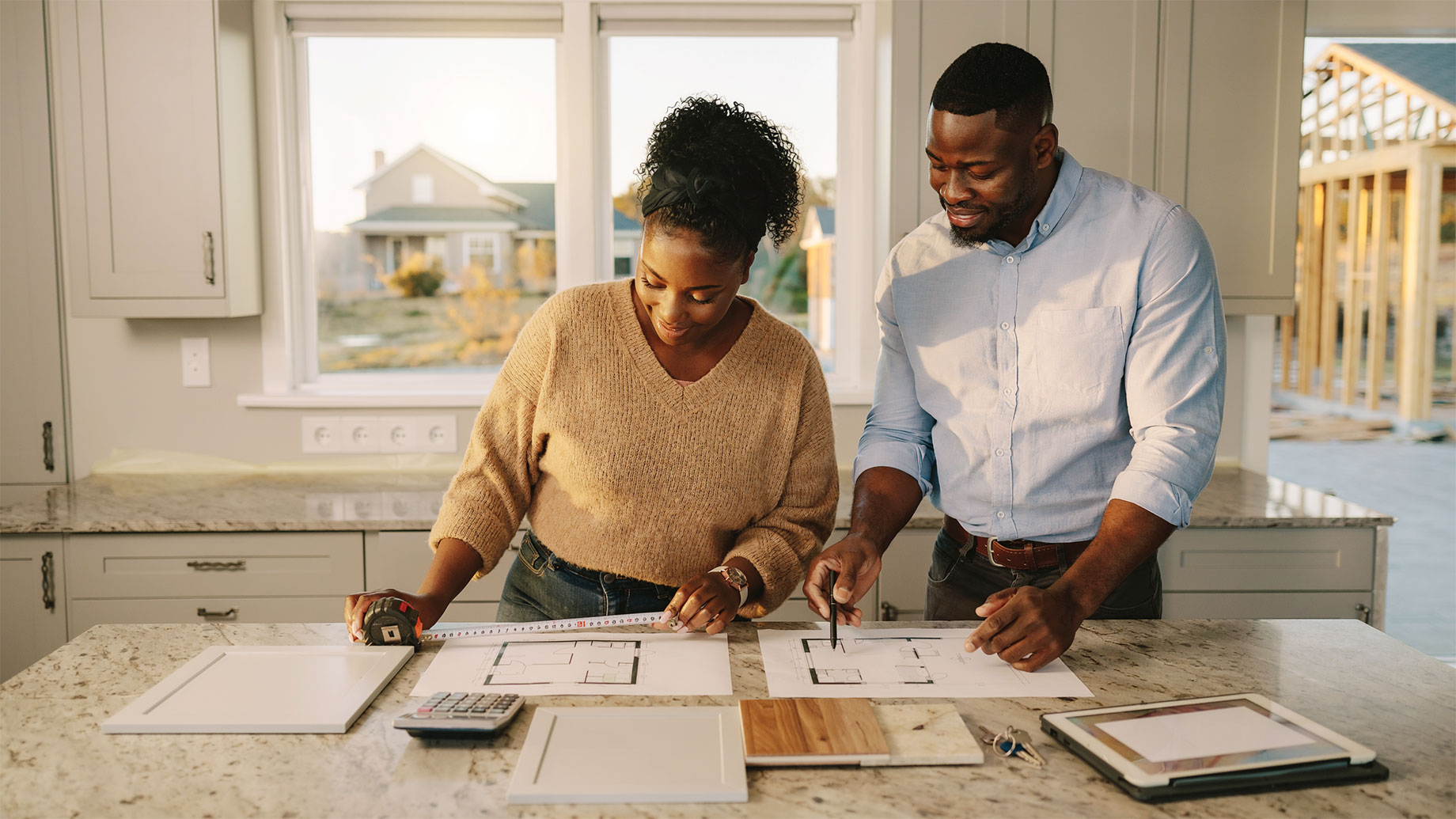 Prospective buyers measure floor plans and compare cabinet and flooring samples in the kitchen of a newly constructed home.