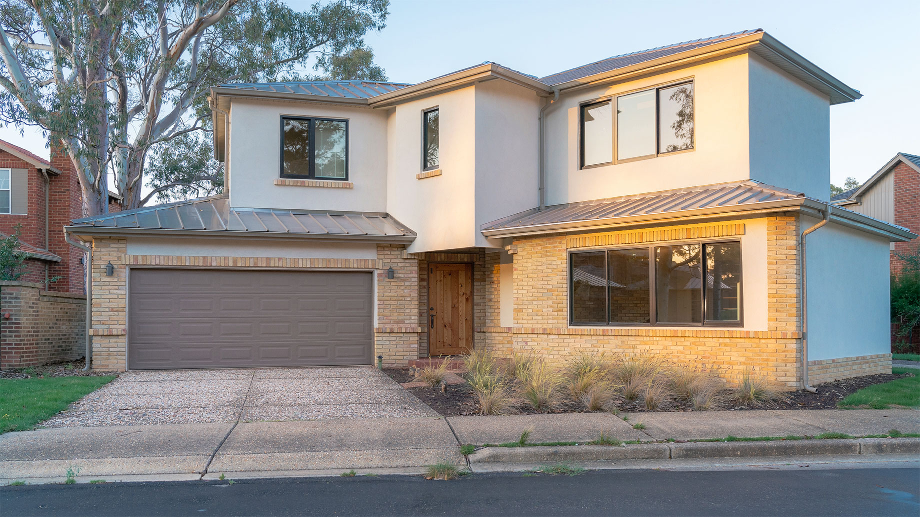 Two-storey home with pale brickwork, white render, a double garage and a eucalyptus tree in warm afternoon light.