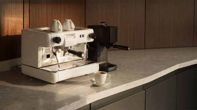 White espresso machine and black grinder on a pale stone counter with an espresso cup, walnut panels, and charcoal cabinetry.