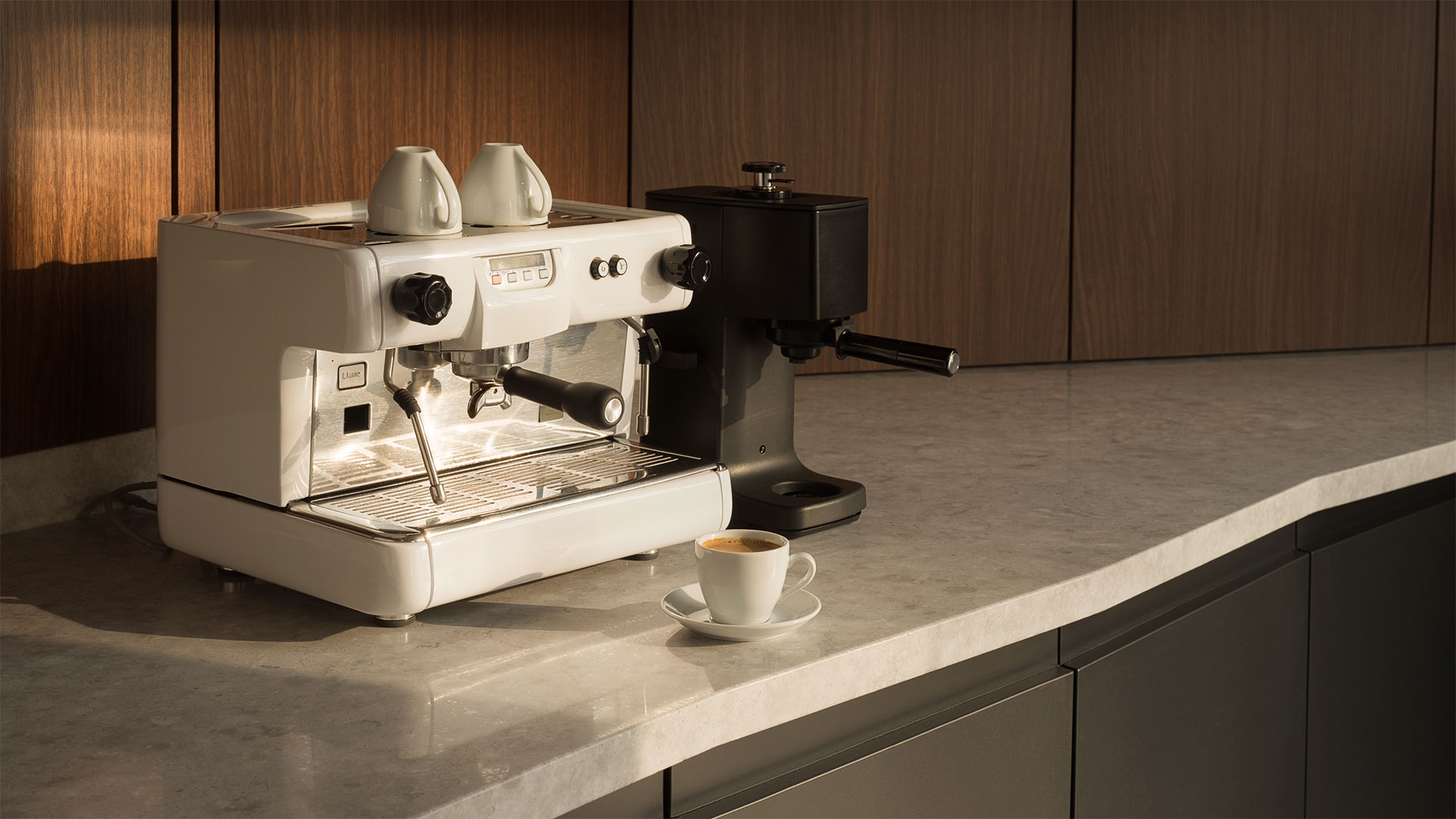 White espresso machine and black grinder on a pale stone counter with an espresso cup, walnut panels, and charcoal cabinetry.