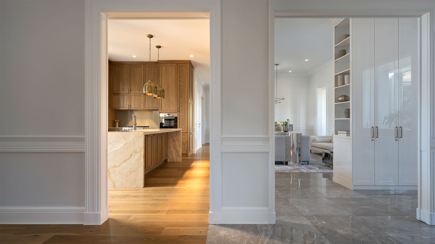 Warm oak kitchen with brass lighting opens into a cool white living area with gray stone floors, revealing a sharp change in finishes between rooms.