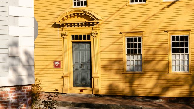 Dark gray paneled door with ornate yellow trim on a yellow clapboard building in Portsmouth, New Hampshire, with tree shadows.