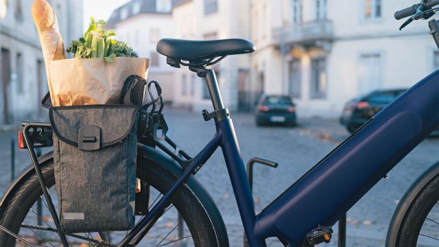 Blue electric bike with a gray rear pannier holding a baguette and vegetables, parked on a cobbled street with cars behind.