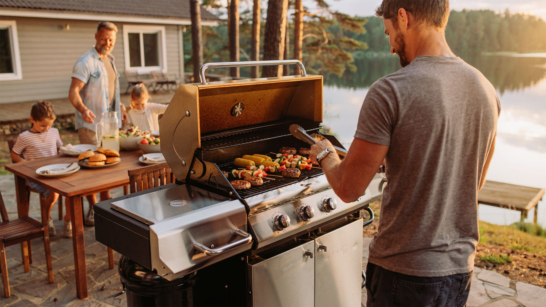 Adult cooking burgers, corn, and vegetable skewers on a propane grill while a family prepares an outdoor meal beside a lakeside cabin.