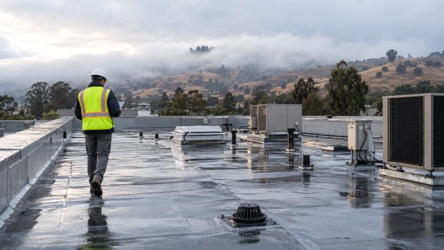 Roof inspector seen from behind on a wet commercial rooftop with HVAC units, vent pipes, and a drain beneath fog-covered hills.