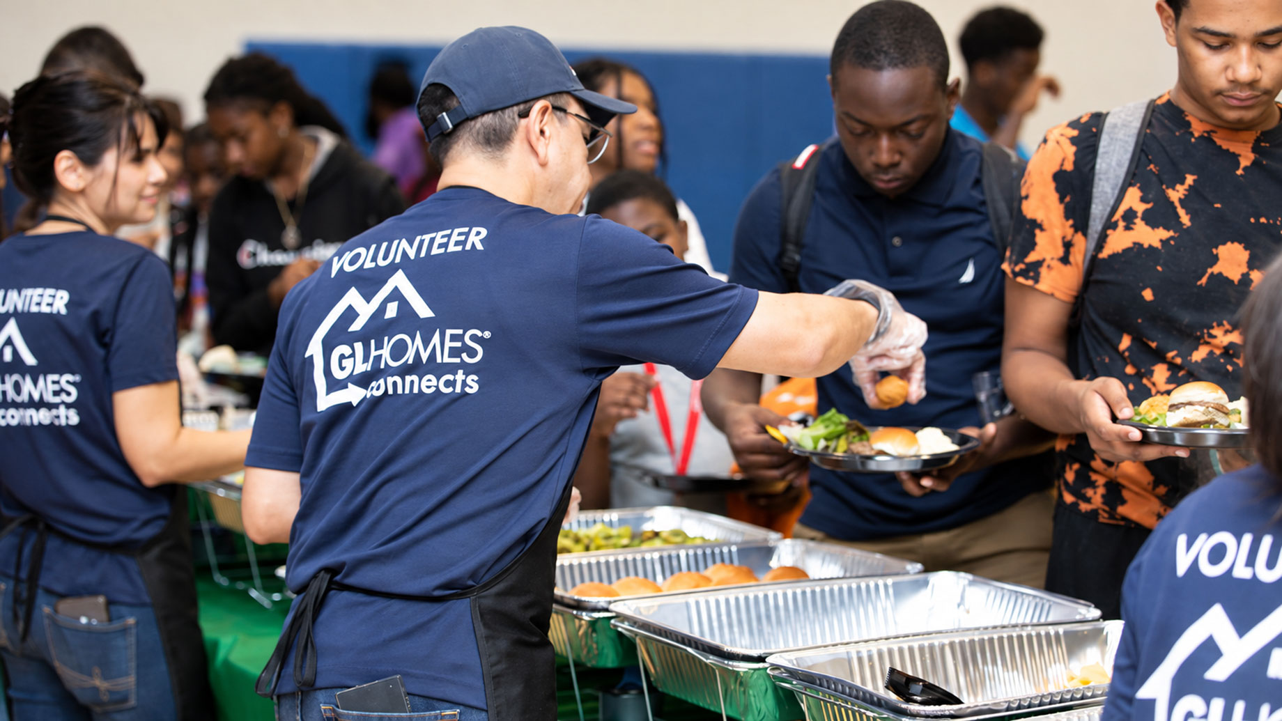 Volunteers wearing GL Homes Connects shirts serve meals from aluminum trays at an indoor community event.