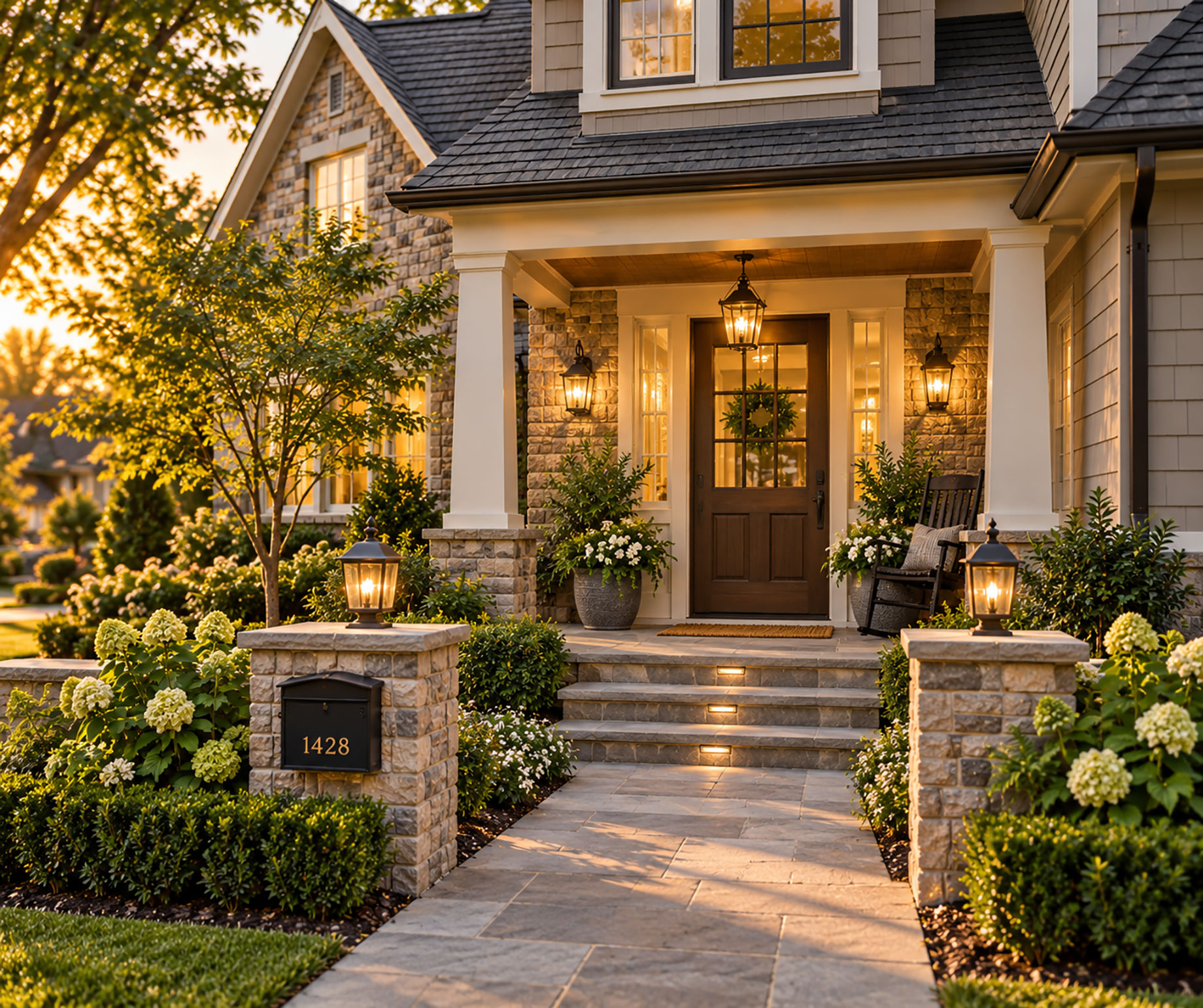 Stone walkway leading to a covered front porch with a wooden entrance door, warm lantern lighting, and landscaped gardens at sunset.