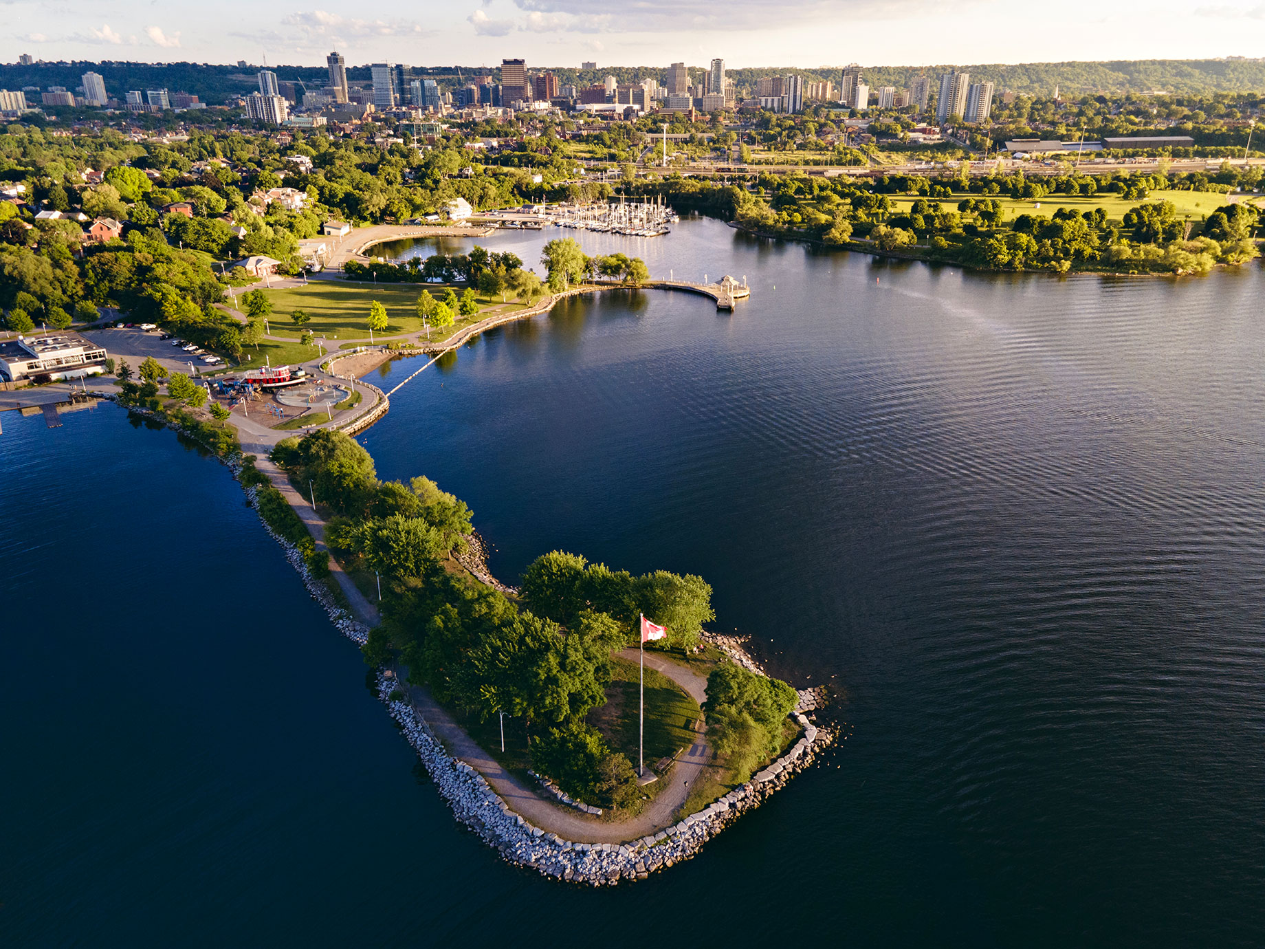 Aerial view of Hamilton, Ontario, showing waterfront parkland, curving shoreline paths, a marina, and downtown buildings beyond the trees.