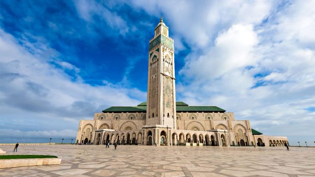 Hassan II Mosque in Casablanca, with an ornate minaret, green-tiled roofs and a patterned courtyard beneath a cloud-filled blue sky.