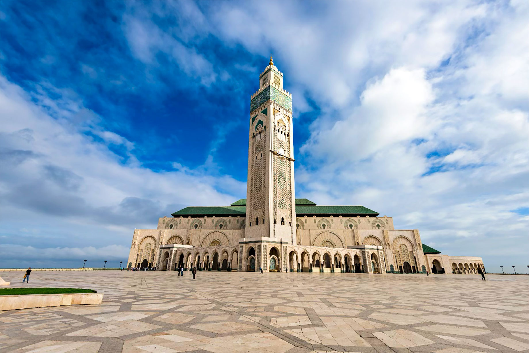 Hassan II Mosque in Casablanca, with an ornate minaret, green-tiled roofs and a patterned courtyard beneath a cloud-filled blue sky.