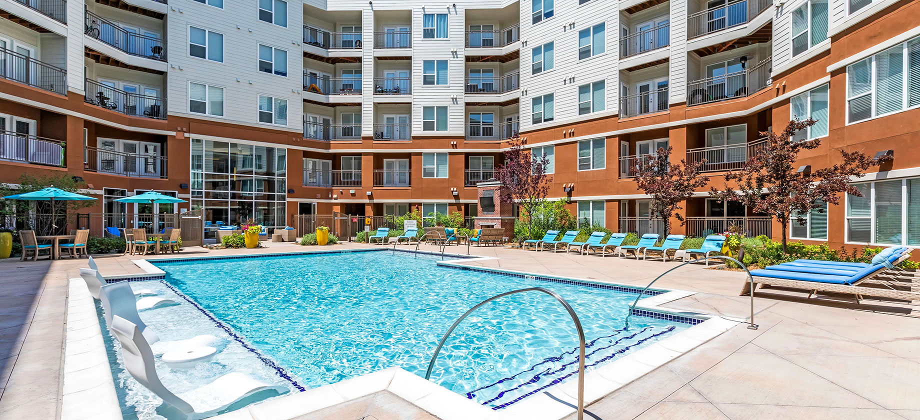 Swimming pool with in-water loungers, blue poolside seating and turquoise umbrellas surrounded by apartment balconies at Helios Apartments.