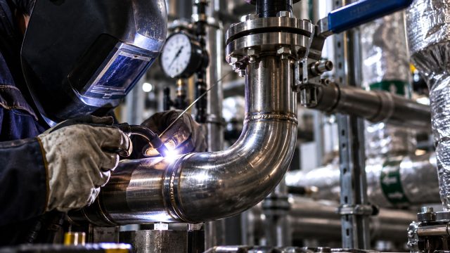 Mechanical contractor wearing a welding helmet and protective gloves welding a stainless steel pipe elbow surrounded by valves, flanges, and industrial equipment.
