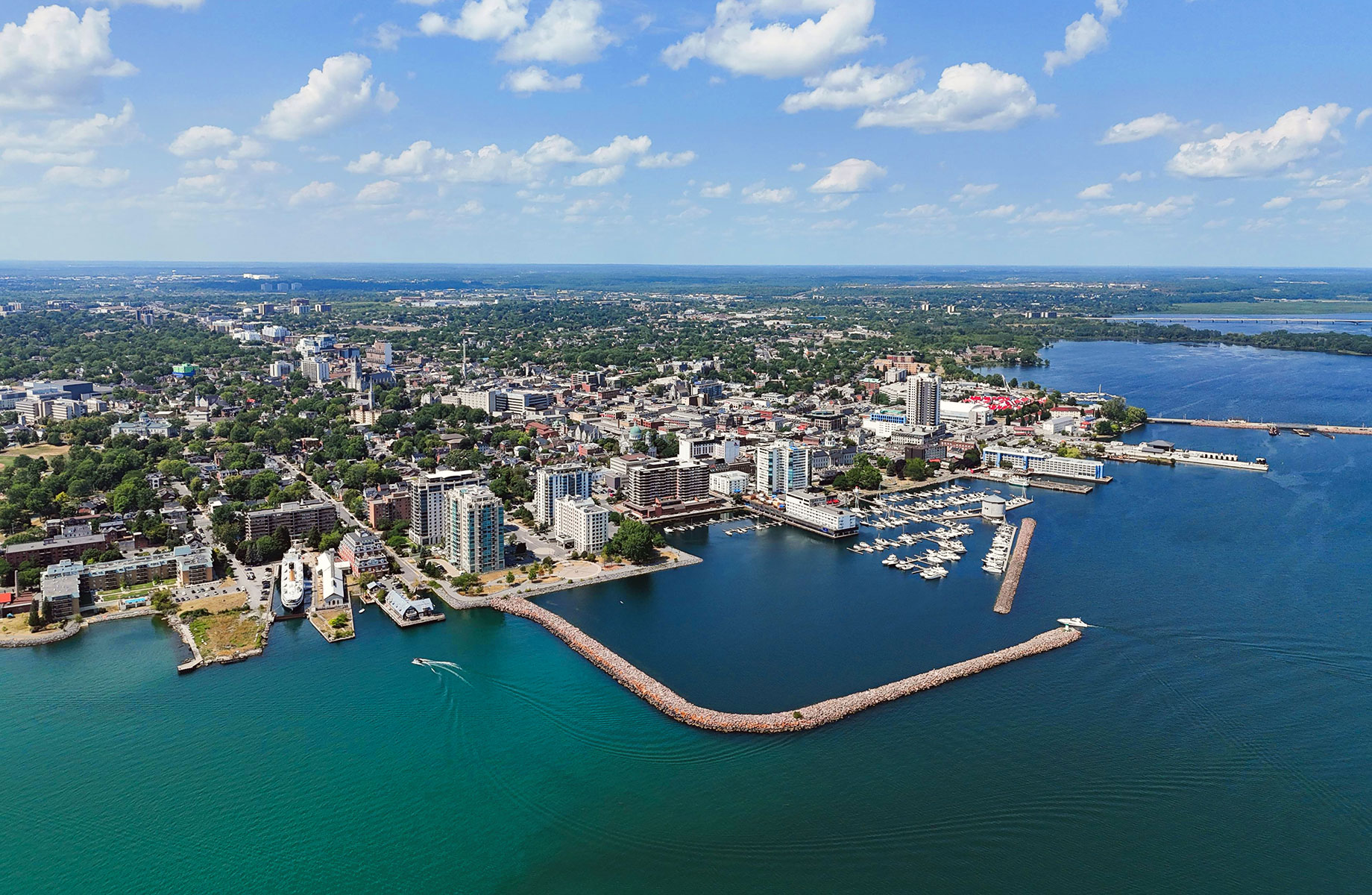 Aerial view of Kingston, Ontario, showing a marina sheltered by stone breakwaters, waterfront buildings, and tree-lined neighbourhoods beside Lake Ontario.