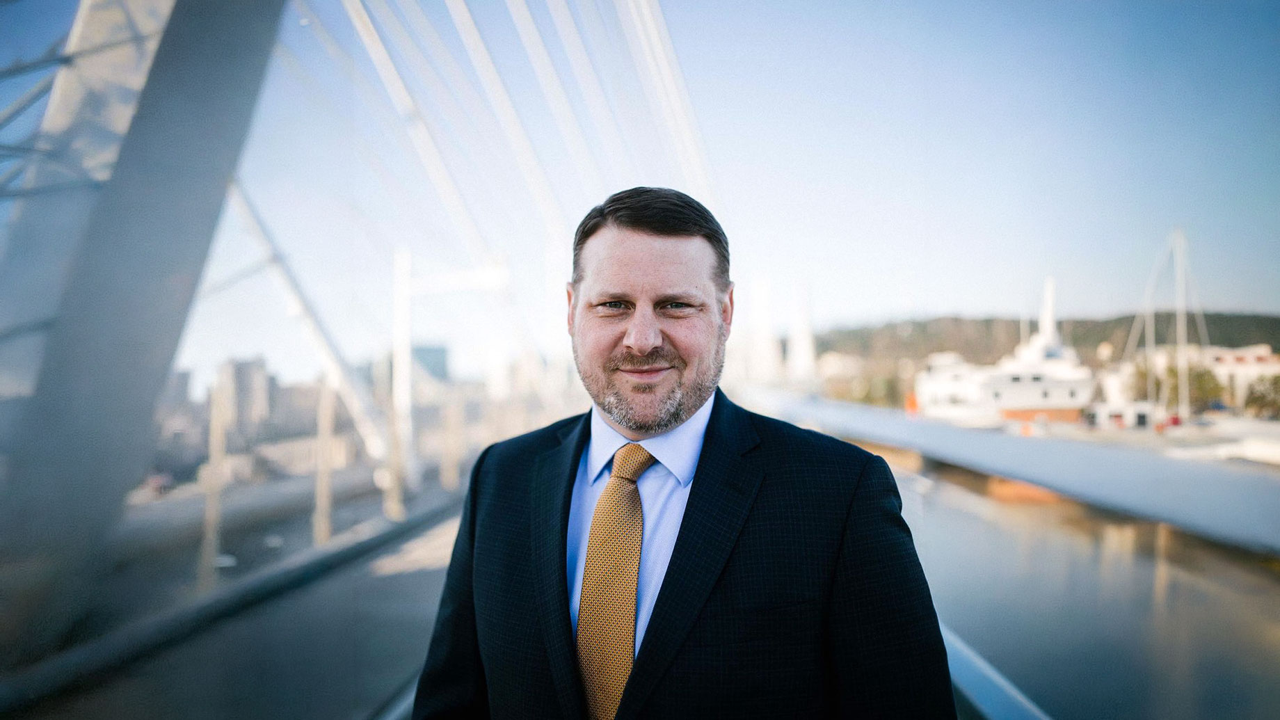 Lewis Irwin Landerholm in a dark suit and gold tie standing on a modern pedestrian bridge with an urban waterfront in the background.