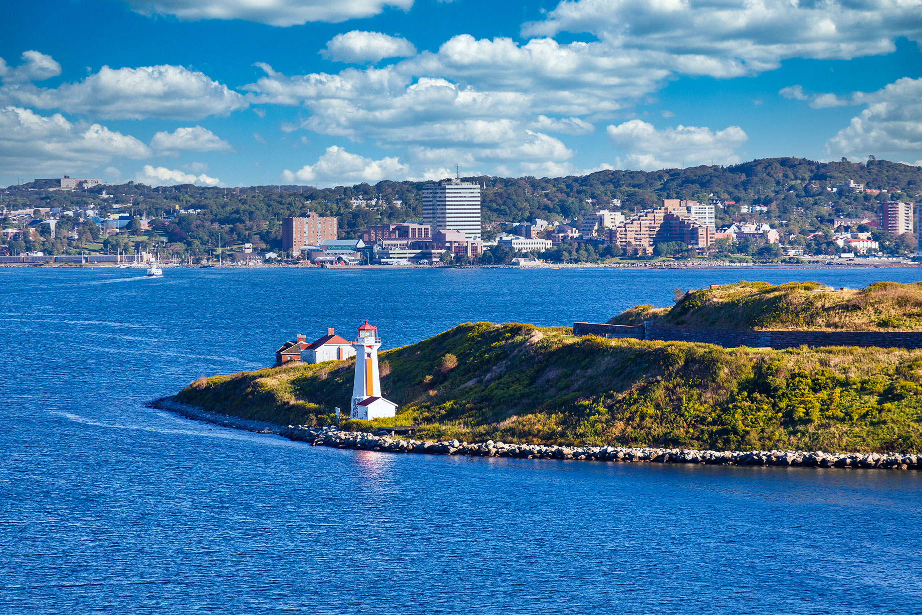Orange-and-white lighthouse on a grassy, rocky shoreline in Halifax Harbour, with waterfront buildings and wooded hills in the distance.