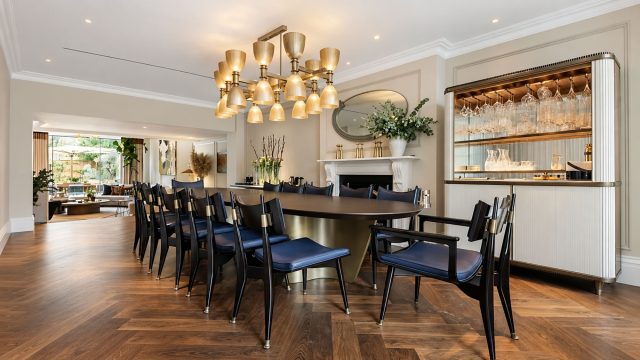 Dining room with a dark oval table, navy chairs, brass chandelier, white fireplace and illuminated glassware cabinet.