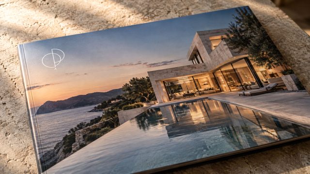 Close-up of a luxury property brochure featuring a coastal villa, infinity pool, and white geometric logo on a textured stone surface.