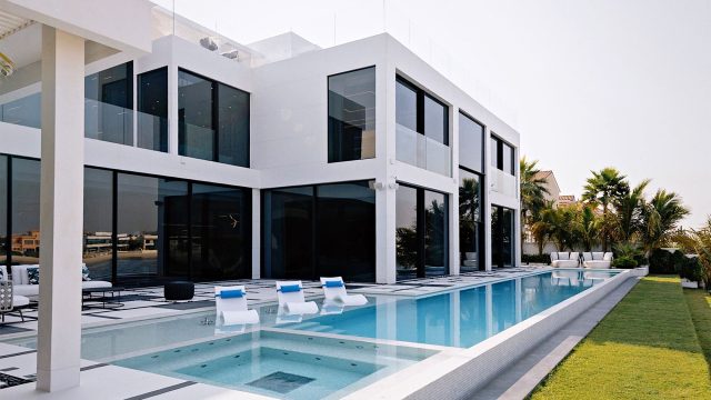 Modern white villa with floor-to-ceiling windows, a swimming pool, three loungers in shallow water and palm trees.