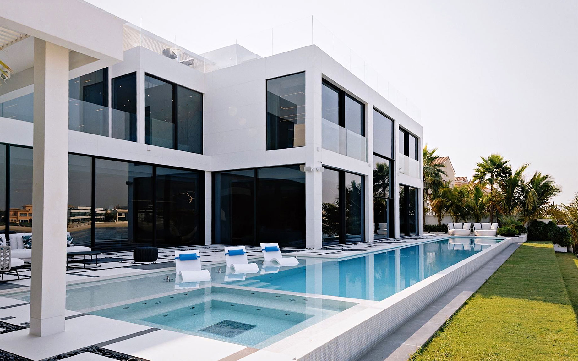 Modern white villa with floor-to-ceiling windows, a swimming pool, three loungers in shallow water and palm trees.