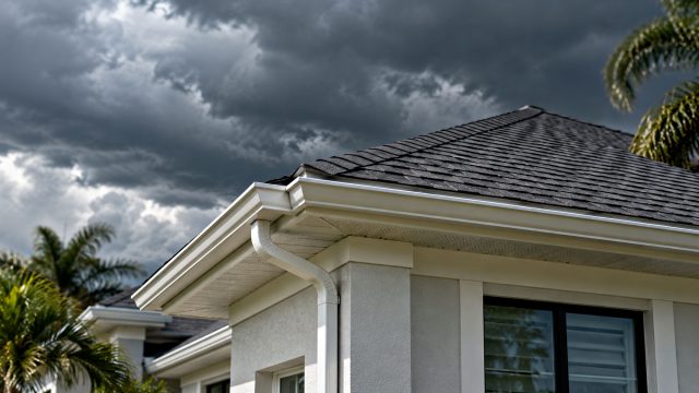 Dark shingle roof with white gutters, a downspout, and vented soffits on a stucco home beneath storm clouds and palm fronds.