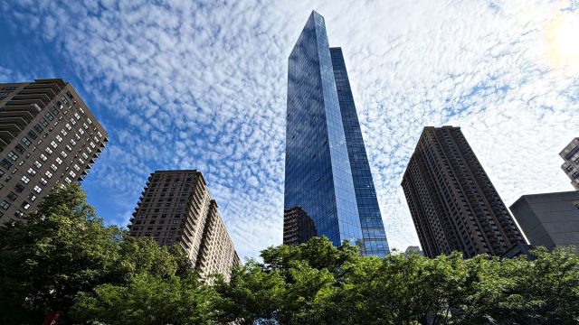 Blue-glass skyscraper between brown masonry apartment towers, viewed from below with leafy trees and white clouds.