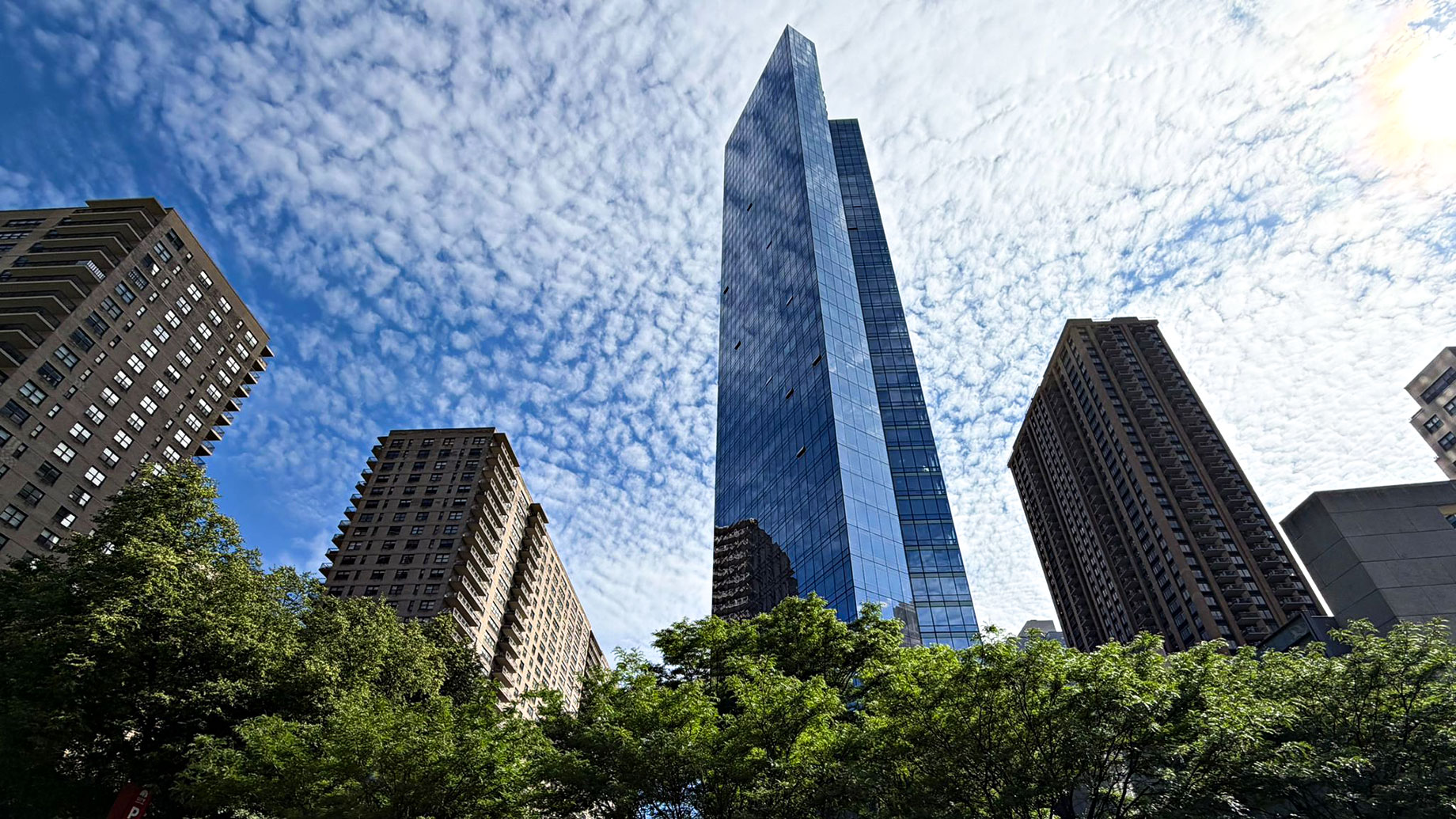 Blue-glass skyscraper between brown masonry apartment towers, viewed from below with leafy trees and white clouds.