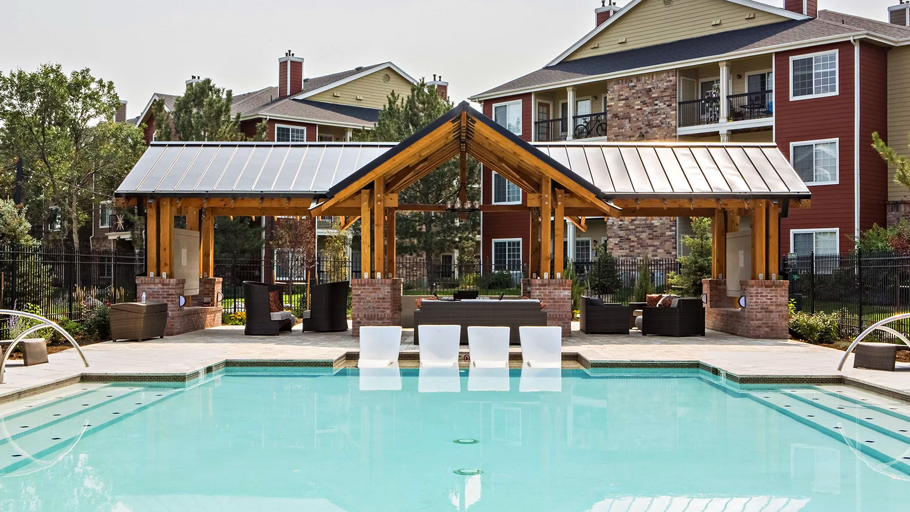 Swimming pool with four in-water loungers and a timber pavilion in front of apartment buildings at Oak Trail at Cherry Creek South.
