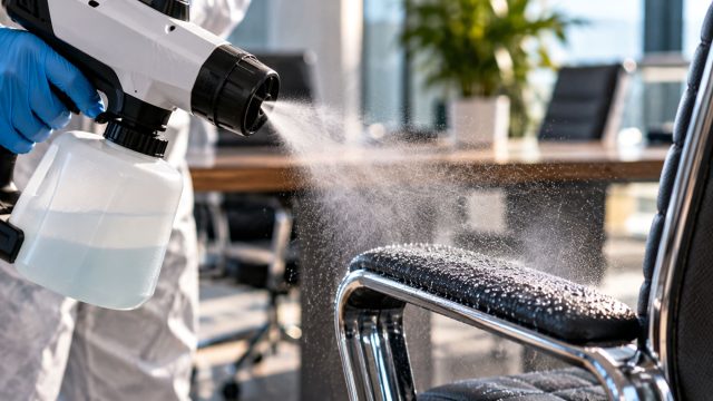 Blue-gloved cleaning technician using a handheld electrostatic sprayer to apply a fine disinfectant mist to a black office chair armrest.