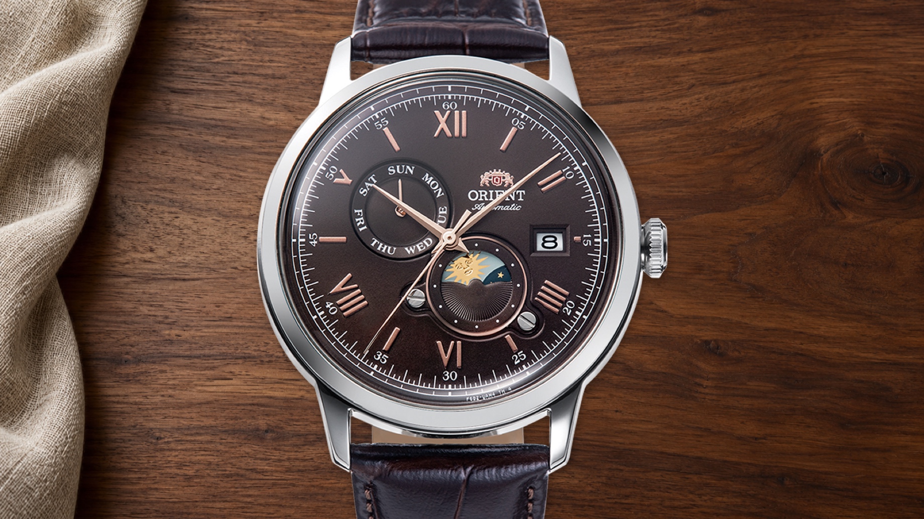 Orient RA-AK0804Y watch with a brown dial, day-and-night display, polished steel case and leather strap against walnut.