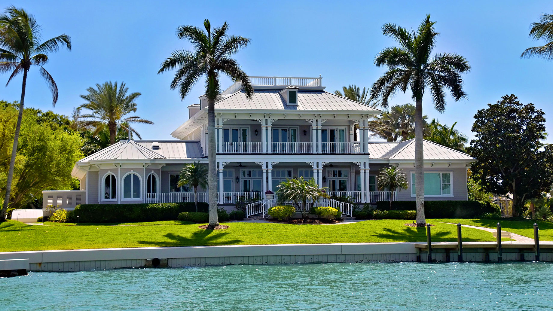 Waterfront home with white-railed porches, a metal roof, palm trees and a manicured lawn beside turquoise water.