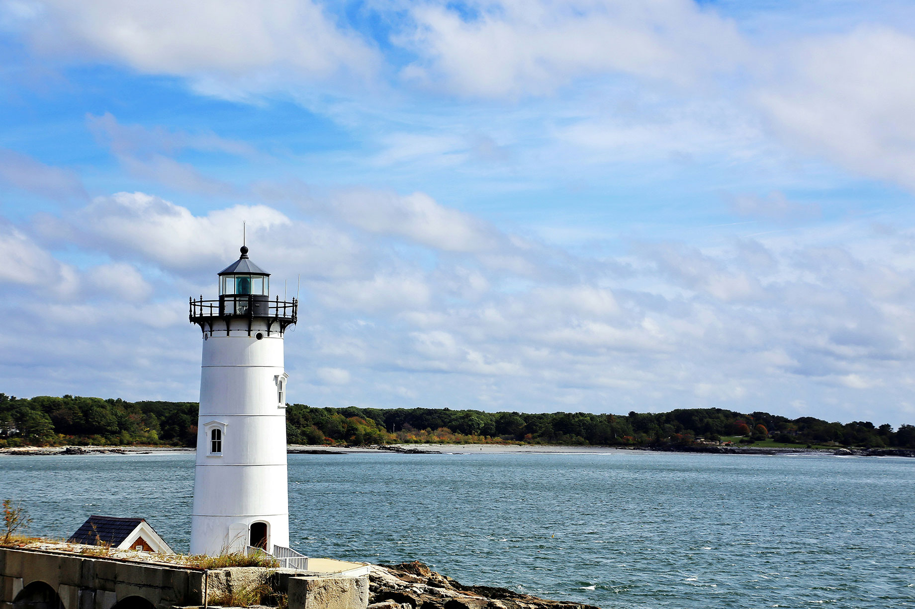 Portsmouth Harbor Light’s white tower and black lantern beside blue water, with a wooded shoreline and clouds behind.