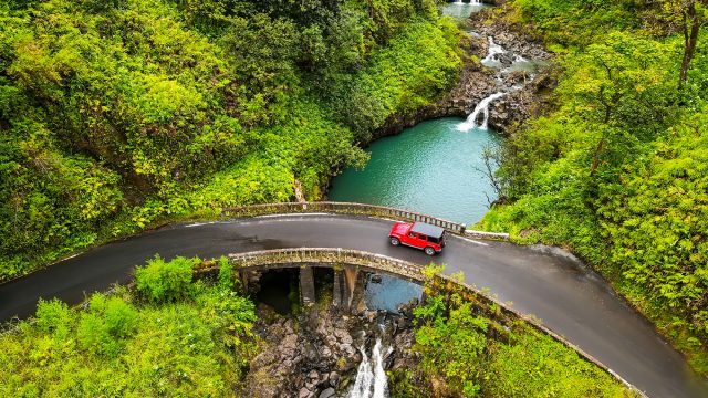 Aerial view of a red SUV crossing a curved bridge above turquoise water and waterfalls surrounded by dense tropical rainforest.